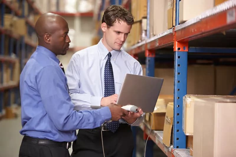 Two warehouse colleagues reviewing stock on a laptop between storage racks